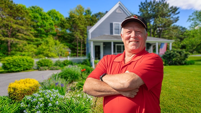 Smiling male handyman in red shirt standing with arms crossed in front of residential home with landscaped driveway.